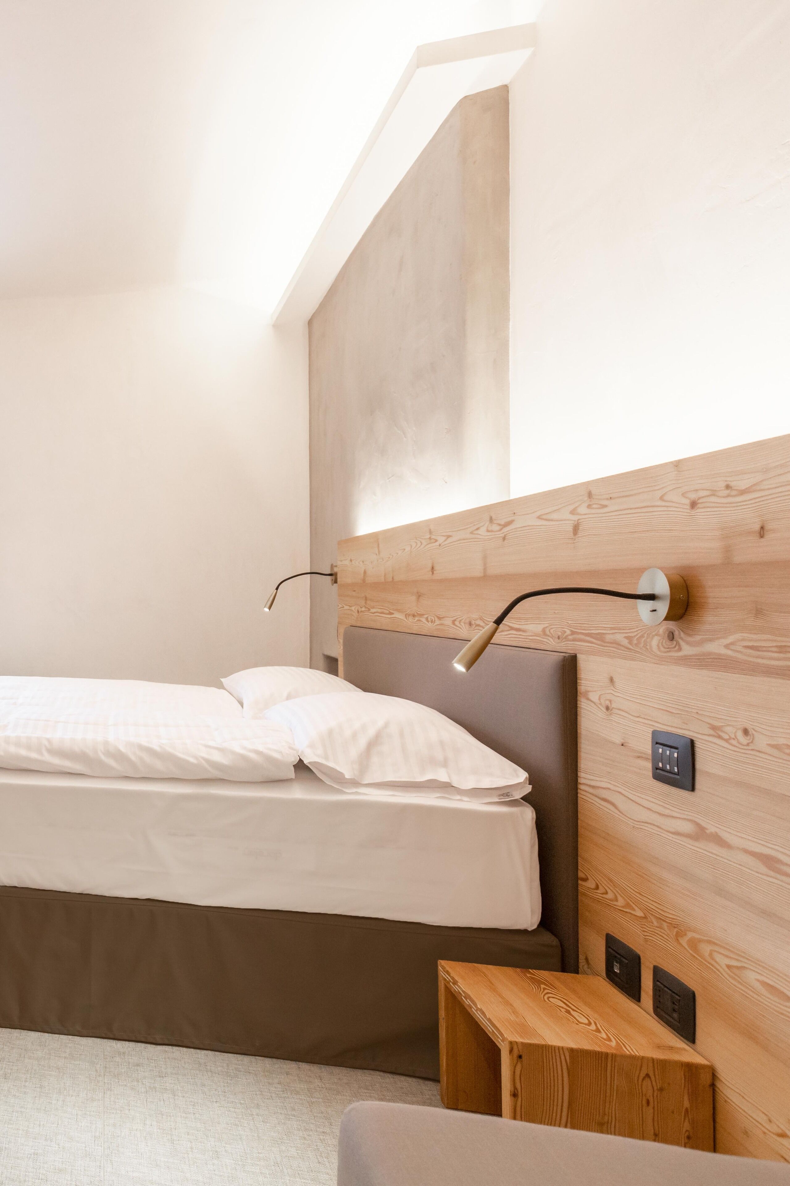 A bedroom with a wooden headboard, a bed with white bedding, a bedside table, and a wall-mounted light fixture.