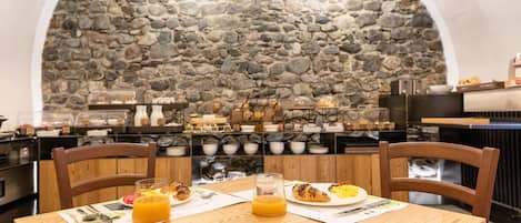 Un desayuno con una pared de piedra de fondo, sillas de madera, una mesa de madera y una variedad de alimentos, incluyendo croissants, fruta y jugo de naranja.