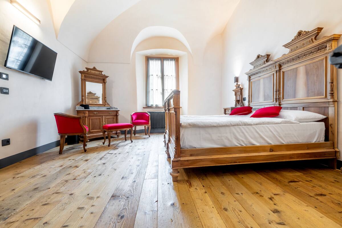 Ein Schlafzimmer mit einem hölzernen Bett, einem roten Sessel, einer Holzkommode, einem Spiegel und einem an der Wand befestigten Fernseher.