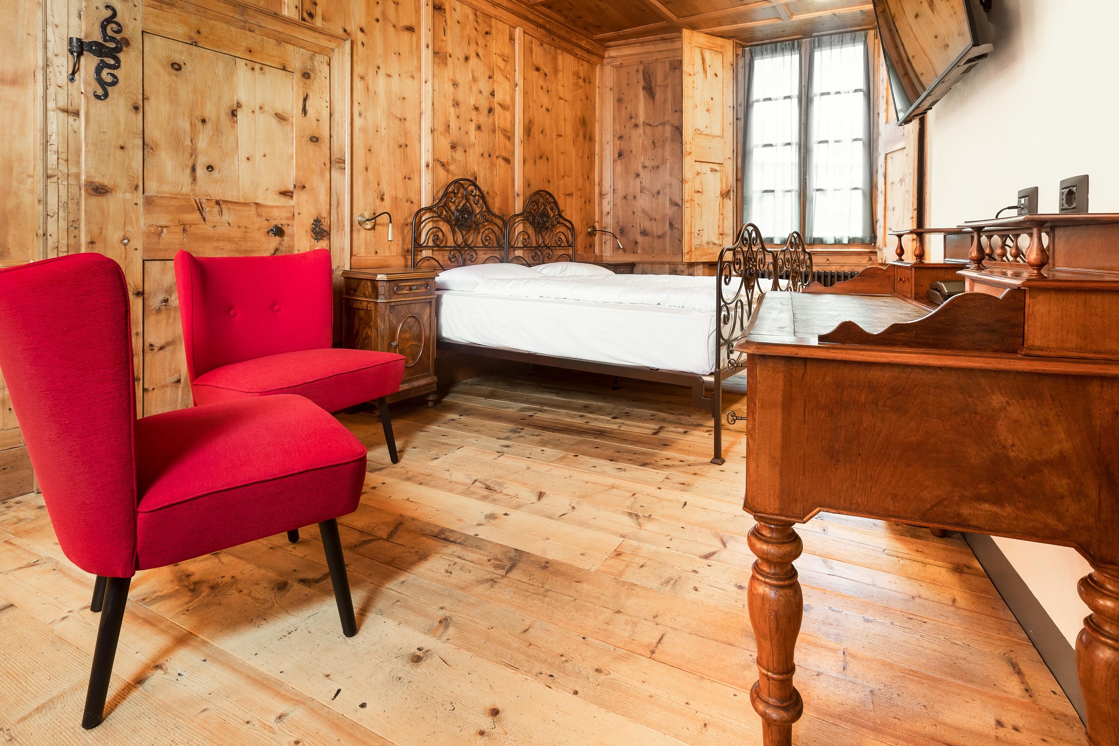 Quarto com painéis de madeira, uma cama, uma poltrona vermelha e uma escrivaninha de madeira.
