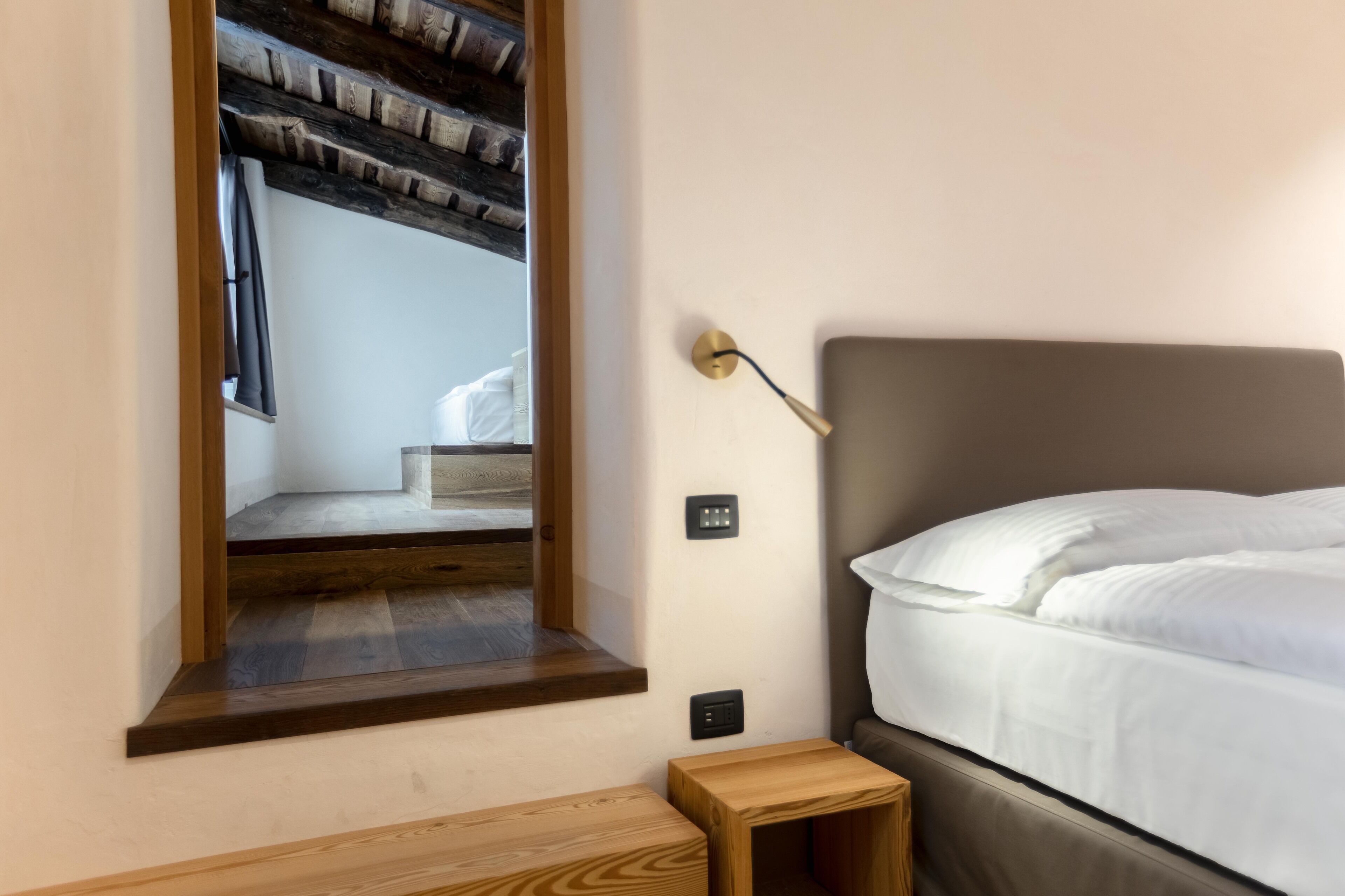 Un dormitorio con cama, una mesita de noche de madera, una lámpara de pared y un espejo que refleja una escalera.