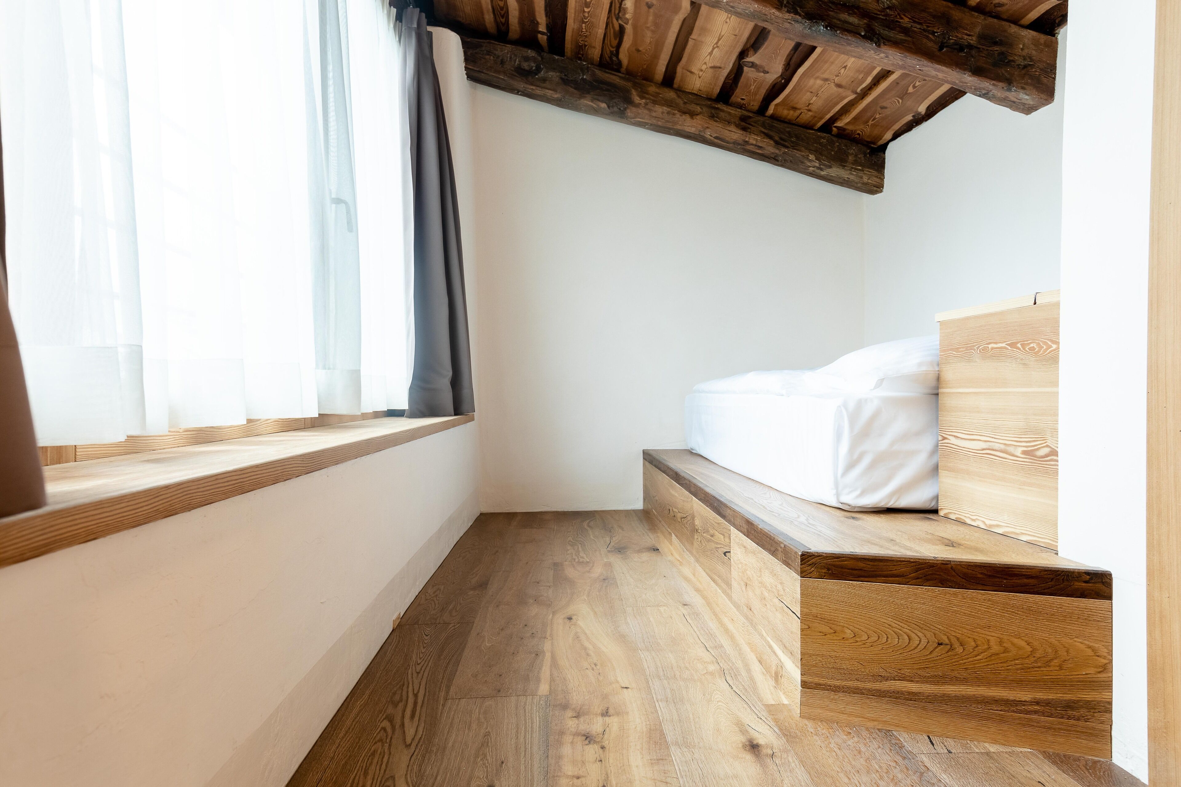 Un dormitorio con piso de madera, una cama con ropa blanca, una ventana con cortinas y un techo de madera.