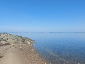 Una costa tranquil·la amb aigua cristal·lina, vorera rocosa i edificis al lluny.