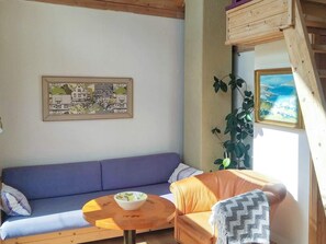 A cozy room with a blue sofa, a round wooden table with a bowl, a checkered armchair, a framed cityscape painting, and a large window with a view of the sky and clouds. - 5 Person Holiday Home in Floda (Floda)