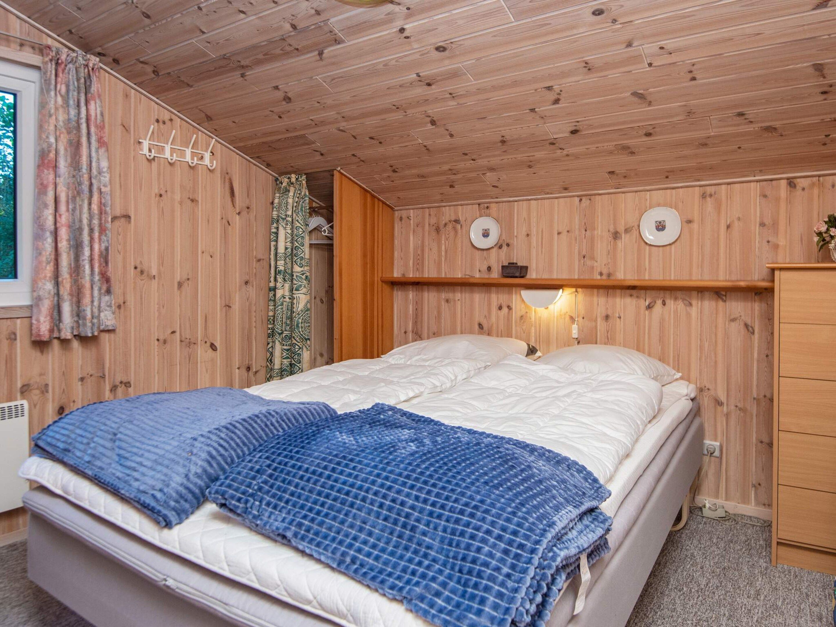 A bedroom with a bed, wooden walls, a window with curtains, and a bedside table with a lamp. - 4 Star Holiday Home in Glesborg (Glesborg)