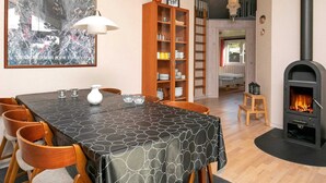 A cozy dining area with a table set for four, surrounded by wooden chairs. A bookshelf filled with books and decorative items is to the right. A fireplace with a black stove is visible in the background.