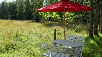 A picnic table with a red umbrella, surrounded by white chairs, set in a grassy field with trees in the background.