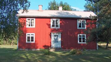 A red two-story house with a white door and windows, surrounded by trees and grass.