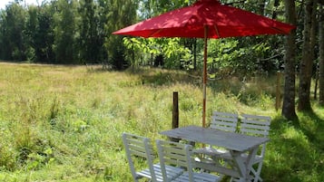 An outdoor seating area with a table and chairs under a red umbrella, surrounded by grass and trees.