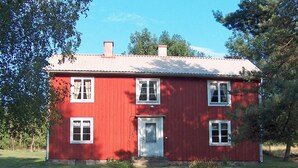 Una casa roja de dos pisos con una puerta y ventanas blancas, rodeada por un patio cubierto de césped y árboles.
