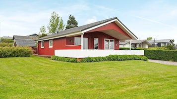 A red single-story house with a white porch, surrounded by a well-maintained lawn and a hedge.