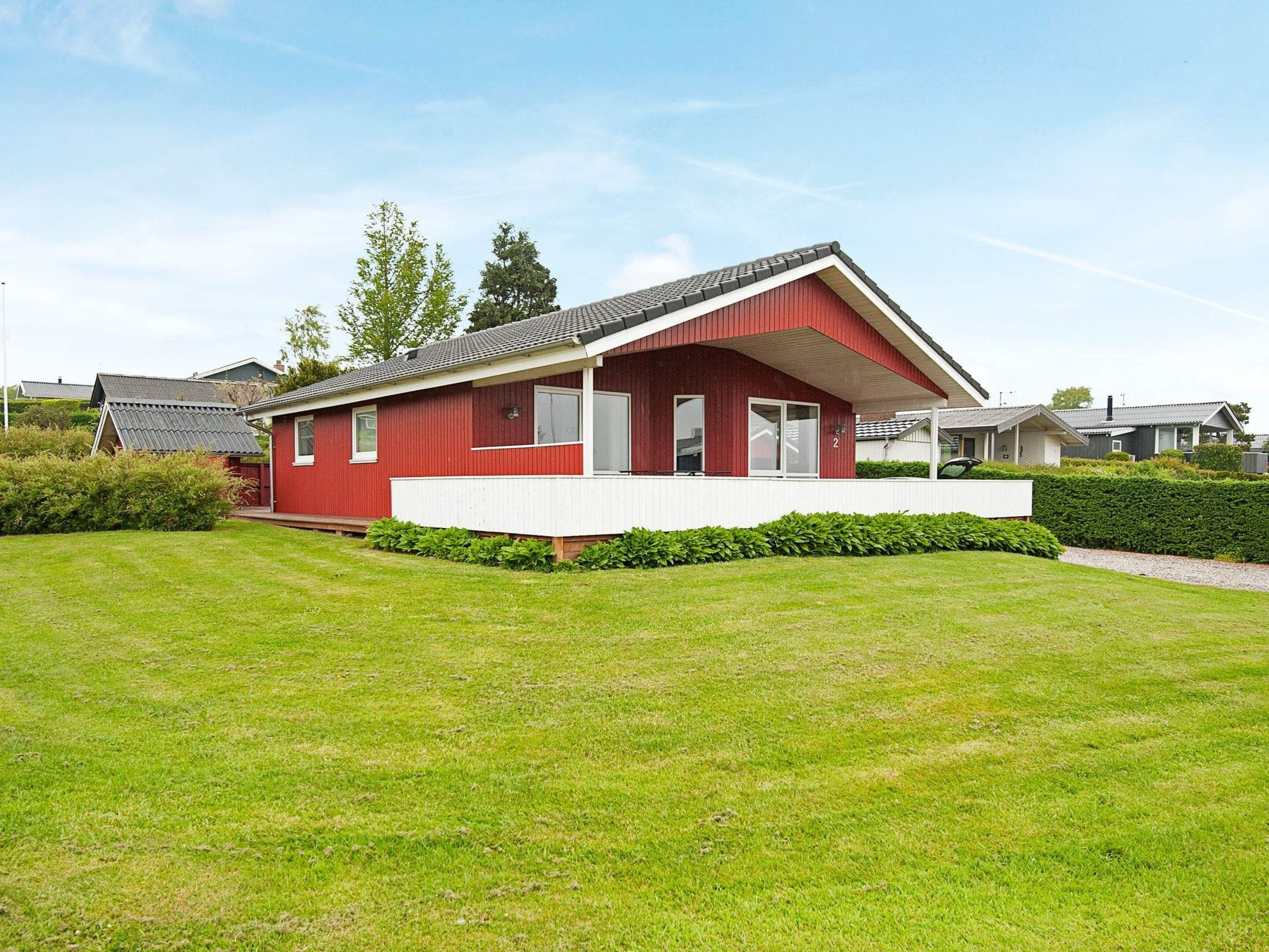 Een rode eenlaagse woning met een witte veranda, omringd door een verzorgd gazon en hagen.
