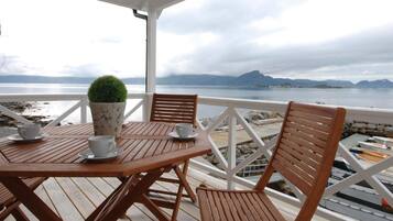 A wooden deck with a table and chairs, a potted plant, and a view of the sea and mountains.