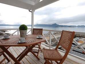 Een houten terras met een tafel, stoelen, een bloempot en uitzicht op zee en bergen.