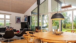 Ein modernes Wohnzimmer mit Esstisch aus Holz, bequemen Sesseln, einem Kamin und großen Fenstern mit Blick ins Freie.