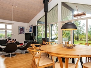 A modern living room with a wooden dining table, comfortable chairs, a fireplace, and large windows offering a view of the outdoors.