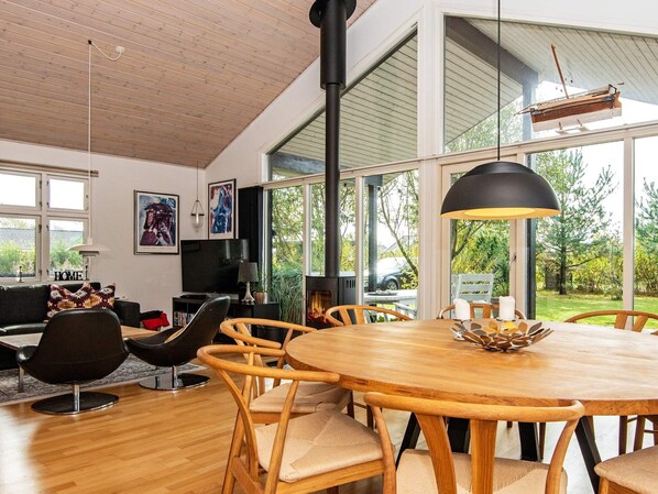 A modern living room with a wooden dining table, comfortable chairs, a fireplace, and large windows offering a view of the outdoors.