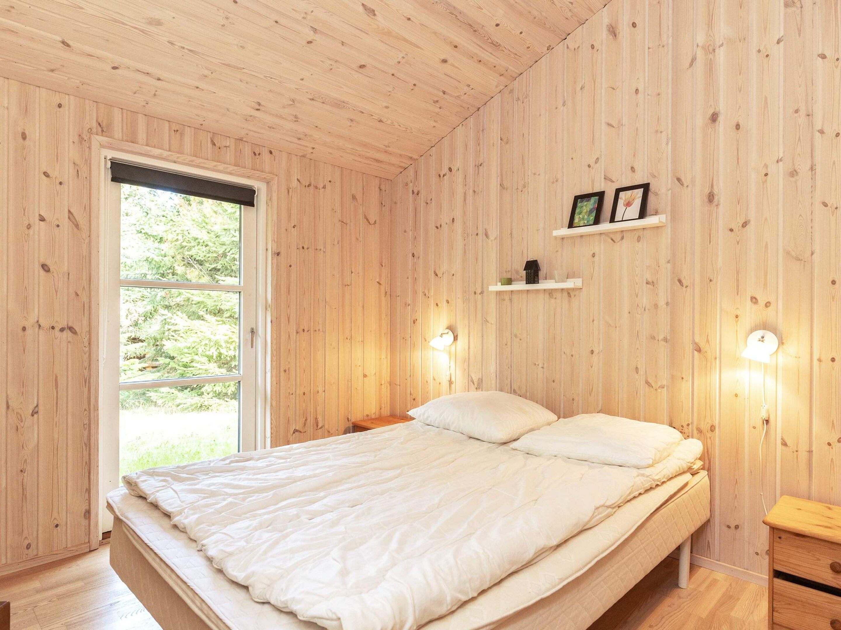 Un dormitorio con piso de madera, cama con ropa de cama blanca, ventana con persianas y dos luces fijadas a la pared.