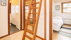 Une échelle en bois dans un couloir menant à une chambre à coucher où se trouvent un lit et une fenêtre.