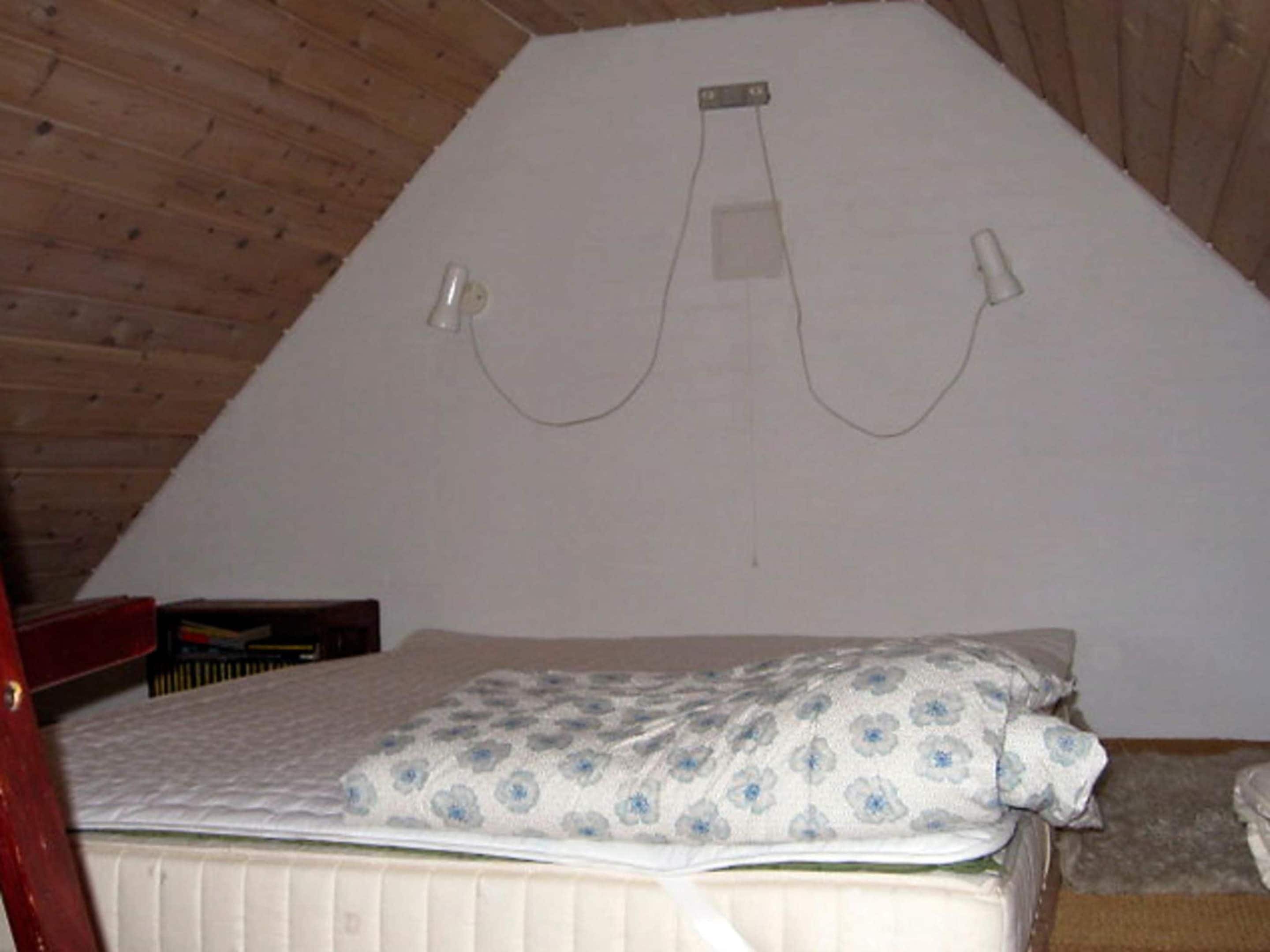 A room with a bed, a wooden ceiling, and a white wall with a simple electrical outlet.