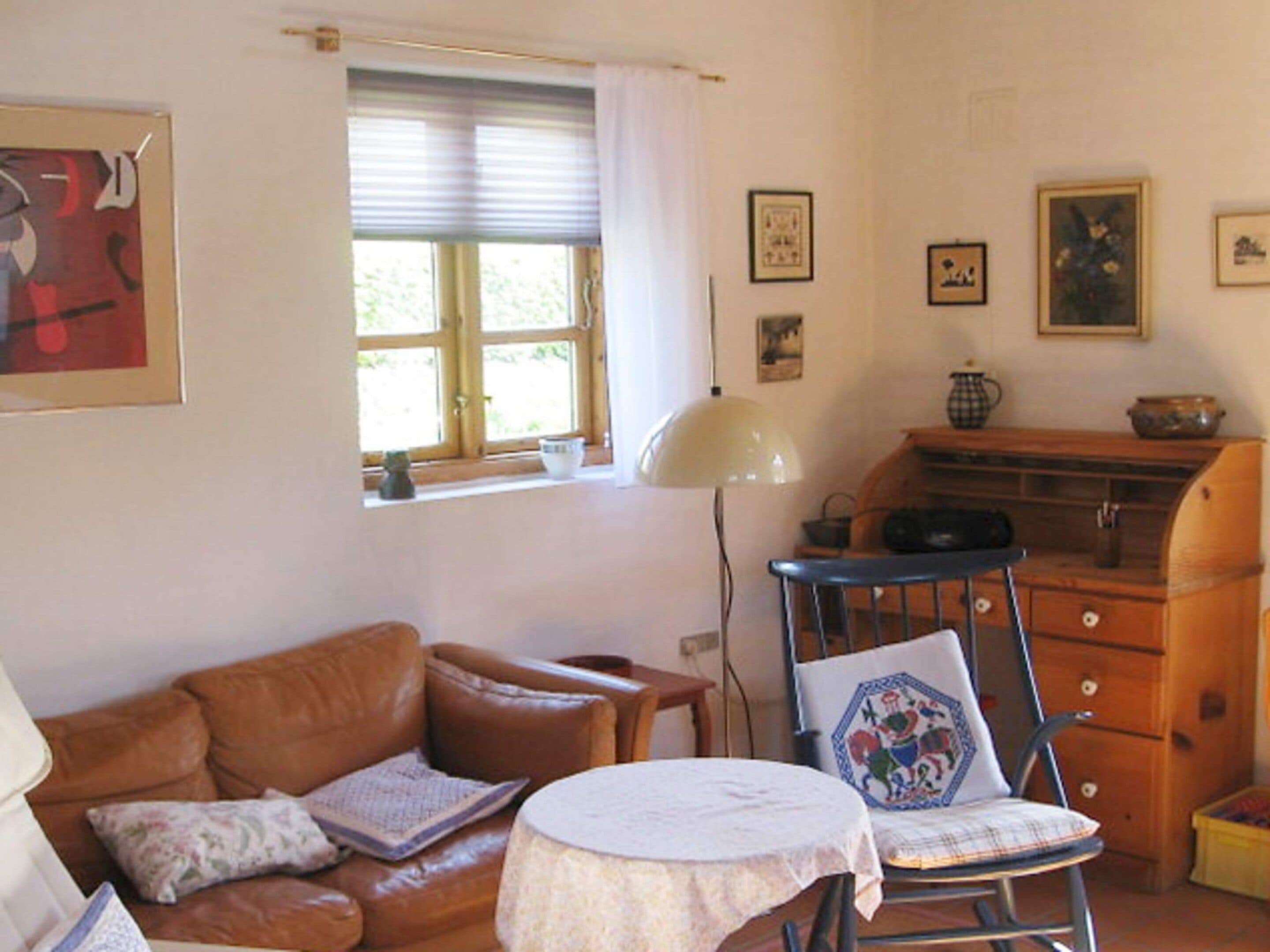 Ein gemütliches Wohnzimmer mit einem braunen Ledersofa, einem kleinen runden Tisch und einem Sessel mit einem Zierkissen. Ein Fenster mit Jalousien und eine Lampe sind zu sehen.