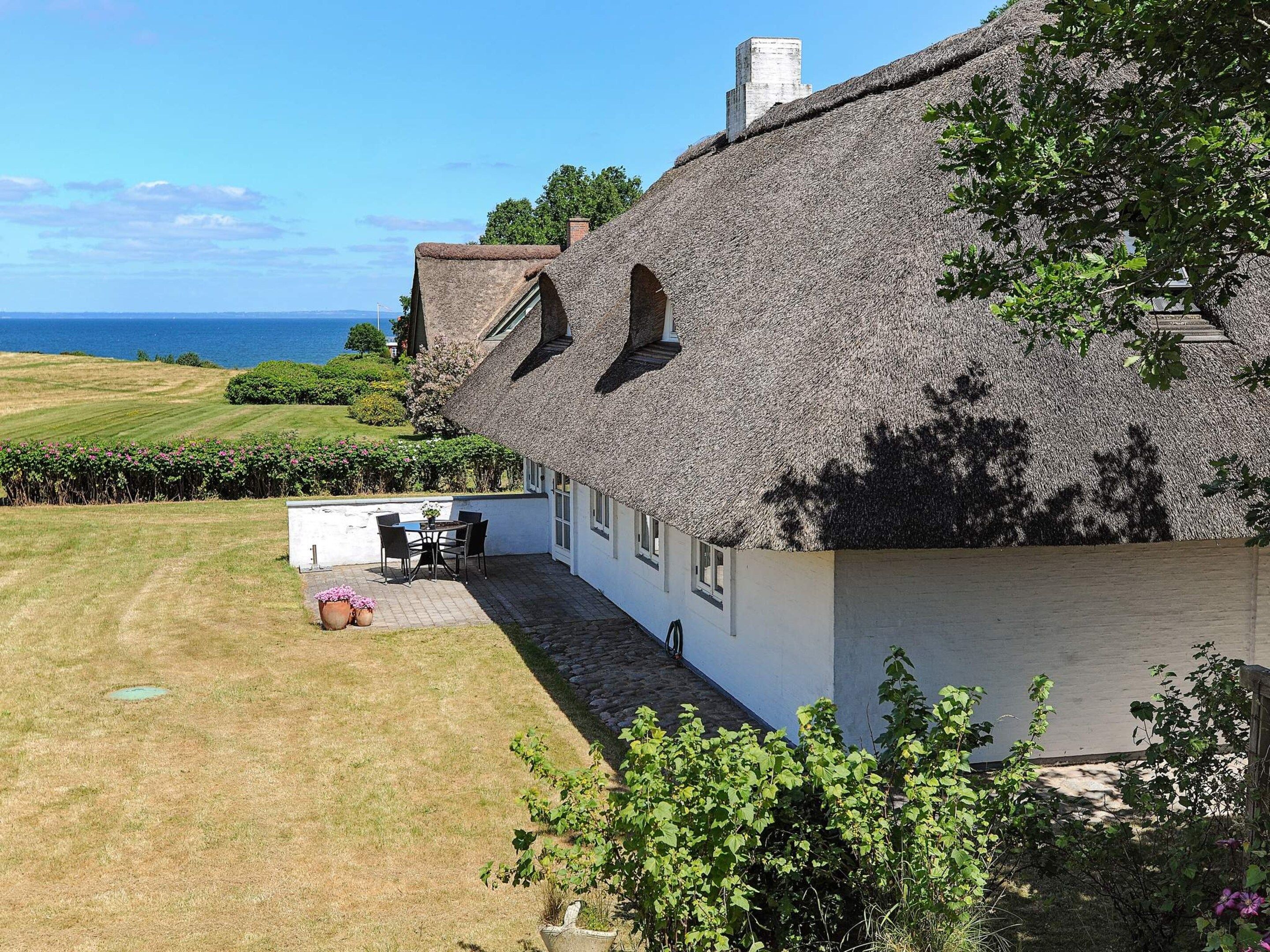 Ein Haus mit Strohdach und Meerblick, eine kleine Terrasse mit Tisch und Stühlen sowie ein Garten mit blühenden Pflanzen.