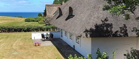 A thatched-roof house with a view of the sea, a small patio with a table and chairs, and a garden with flowering plants.