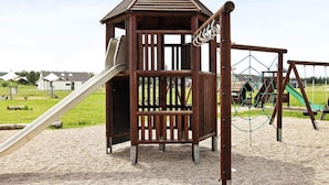 A wooden play structure with a slide and climbing net, set on a sandy playground.