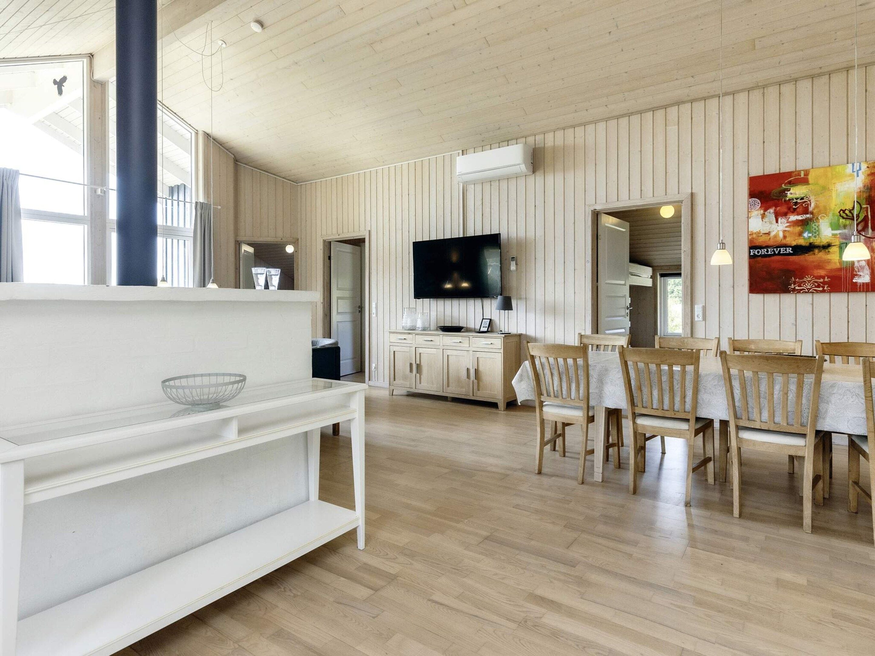 A spacious room with a wooden floor, a dining area with wooden chairs and a table, a television mounted on the wall, and a large window providing natural light.