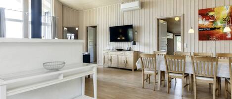 A spacious room with a wooden floor, a dining area with wooden chairs and a table, a television mounted on the wall, and a large window providing natural light.
