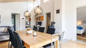 Un espace repas moderne avec une table en bois, des chaises noires et un saladier rempli de fruits. Des luminaires suspendus sont placés au-dessus de la table, et un mur en briques blanches se trouve à l’arrière-plan.