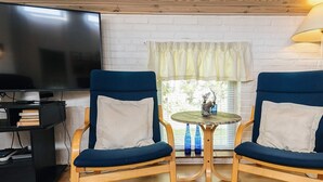 Una sala de estar con dos sillones azules, una mesita redonda, un televisor y un ventanal con cortinas.