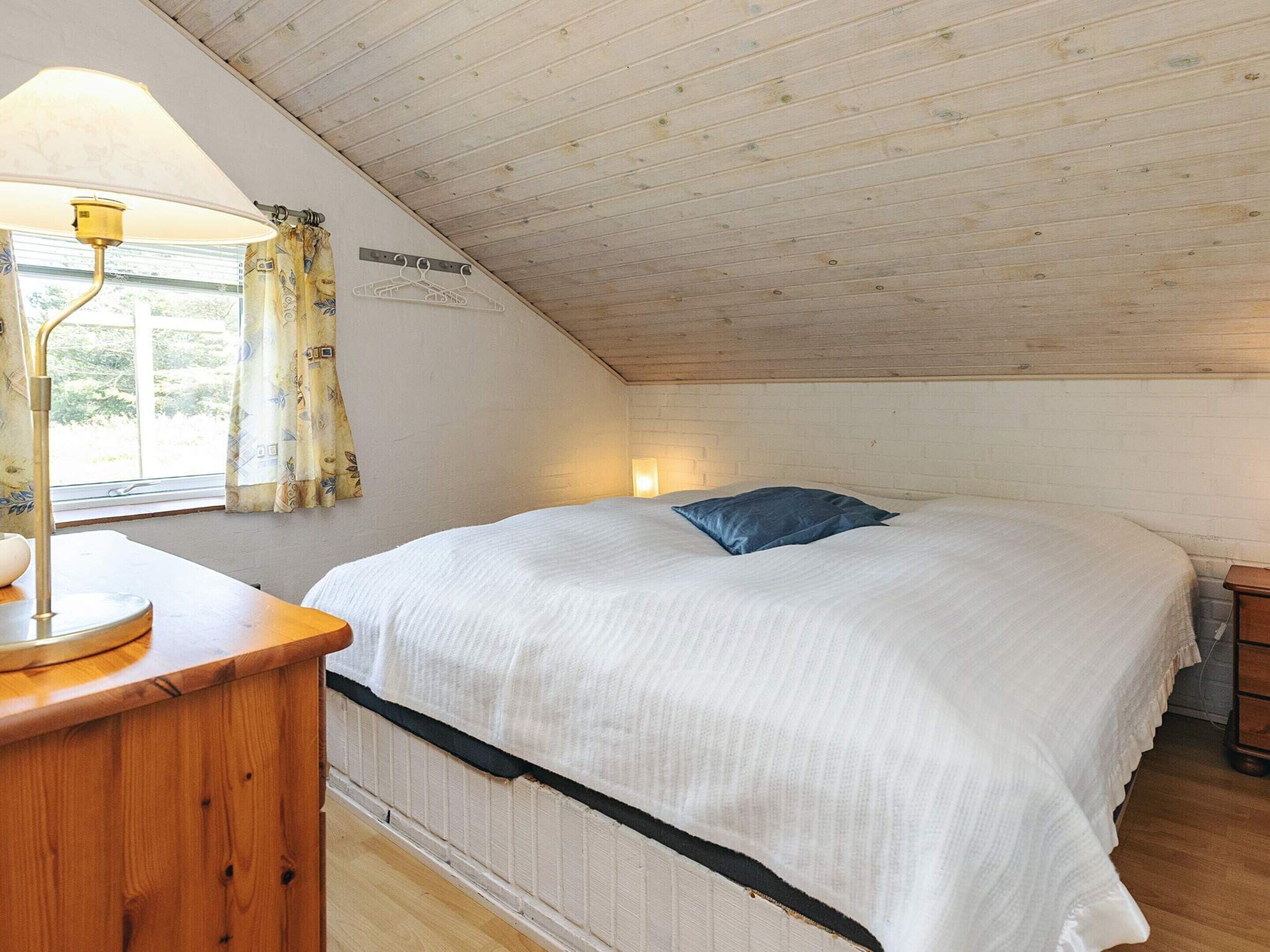 Un dormitorio con techo inclinado, una cama con edredón blanco y almohada azul, una mesita de noche con lámpara y una ventana con cortinas.