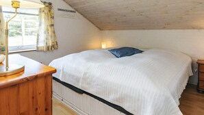 A bedroom with a sloped ceiling, a bed with a white comforter and a blue pillow, a bedside table with a lamp, and a window with curtains. - 7 Person Holiday Home in Thisted (Thisted)
