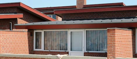 A two-story brick building with a red roof, a visible chimney, and a window with curtains.