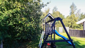 A playground with a swing set and slide, surrounded by a wooden fence and greenery. - Luxury Retreat Near Beach - By Traum Ferienwohnungen (Otterup)