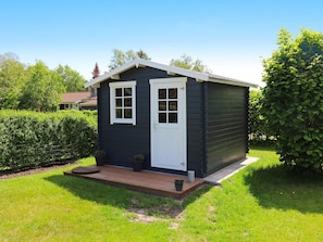 Un petit abri de jardin de couleur sombre, avec une porte et des fenêtres blanches, une terrasse en bois et une pelouse bien entretenue.
