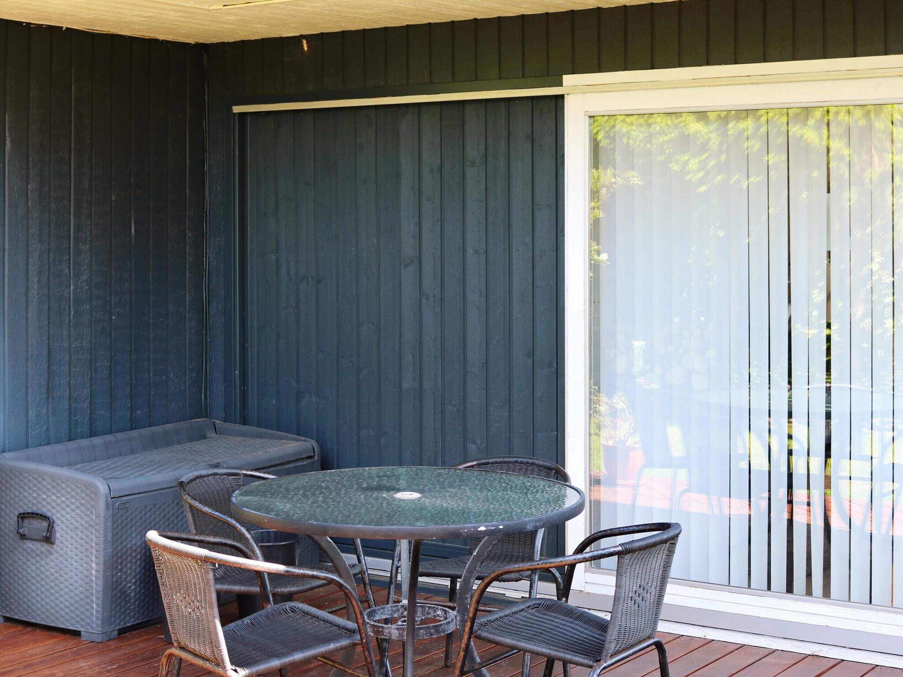 Een overdekt terras met een ronde tafel, vier stoelen, een bank en een schuifdeur van glas.