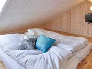 Une chambre avec un plafond en pente, des murs en bois, un lit avec une literie blanche et des oreillers à motifs, et un téléviseur fixé au mur.