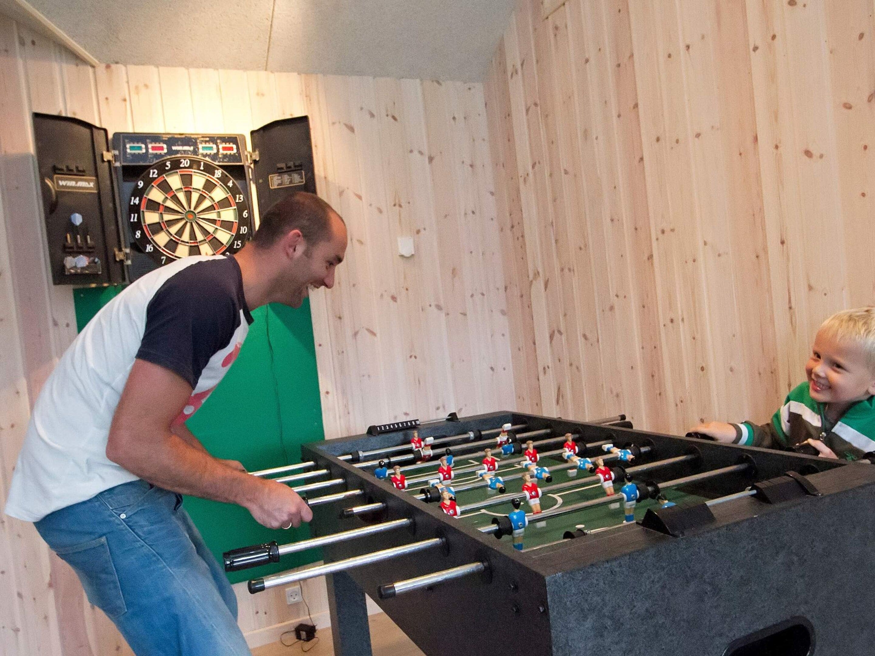 A man playing foosball with a child in a room with wooden walls and a dartboard.