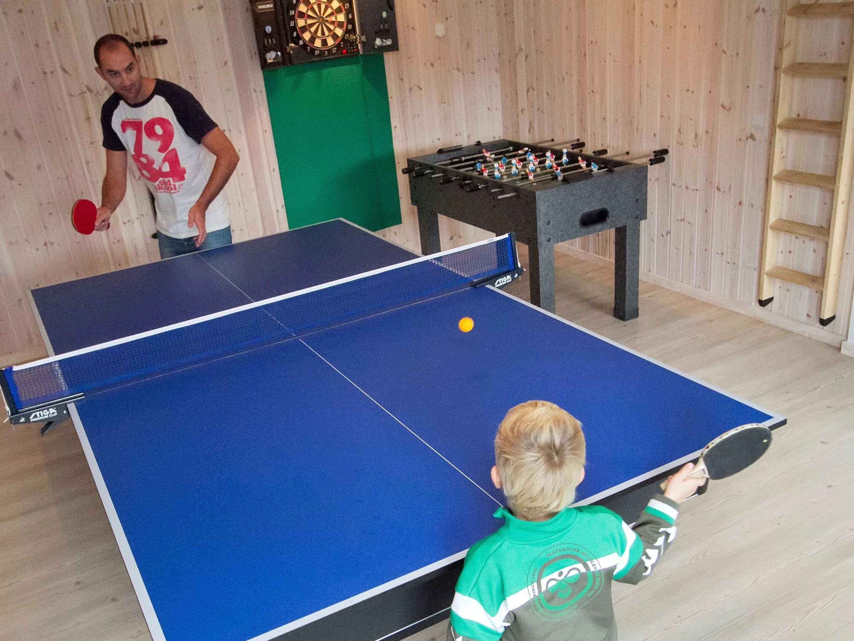 A ping pong table with a net, a foosball table, and a dartboard in a room with wooden walls.