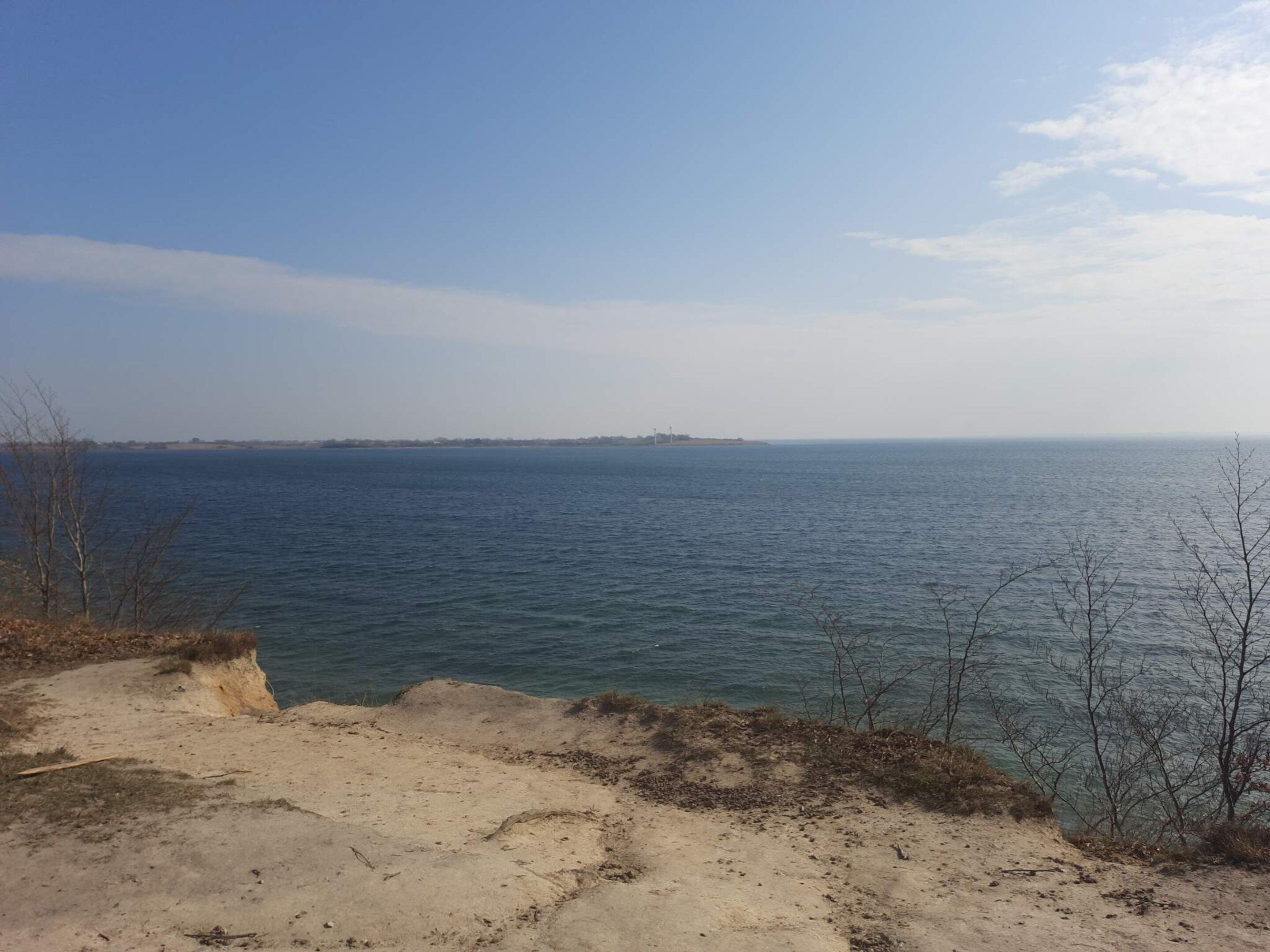 Un paisaje costero con un primer plano arenoso, un mar tranquilo y una costa distante.