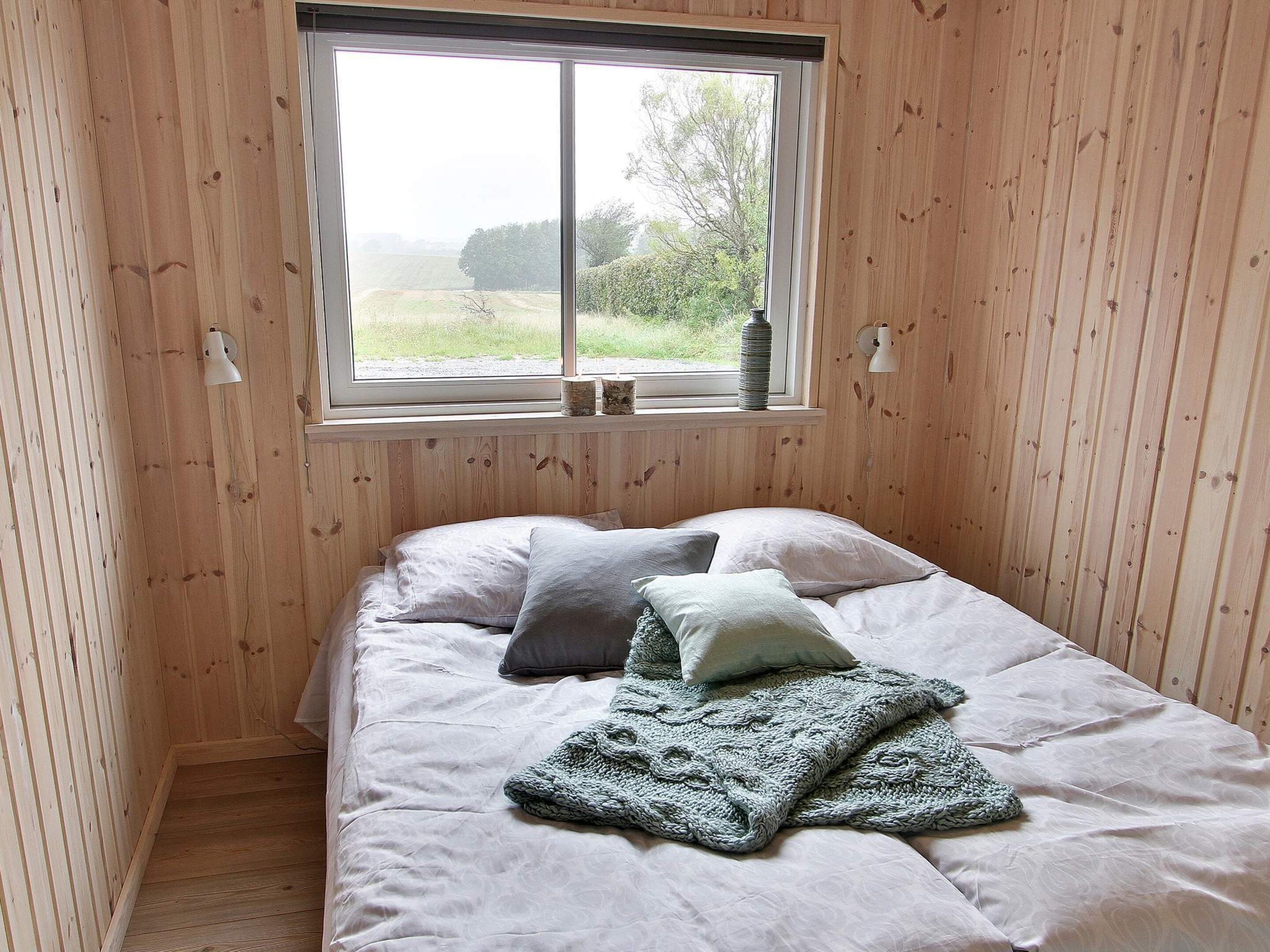 A room with wooden walls, a bed with pillows and a blanket, a window with a view of greenery, and a wall-mounted lamp.