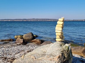 En kustvy med en hög av stenar på en klippa, en strand med småsten och ett lugnt hav.
