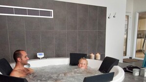Ein Whirlpool mit zwei Personen und einem Kind, umgeben von grauen Fliesen und einer weißen Wand mit Lüftung.