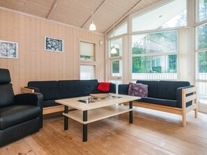 A cozy living room with a wooden ceiling, large windows, a black sofa, a coffee table, and a red throw pillow. - 5 Star Holiday Home in Oksbol (Oksbol)