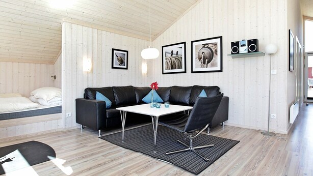 Habitación moderna con un sofá de cuero negro, una mesa blanca y un tapete negro. Se observan cuadros enmarcados en la pared y un altavoz a la derecha.