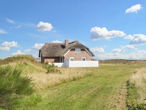 Une maison avec un toit de chaume, des murs en briques et une clôture blanche, située dans un champ verdoyant.