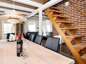 Un coin repas avec une table en bois, des chaises, une bouteille de vin et un verre. On aperçoit un mur en briques et un escalier en bois à l’arrière-plan.
