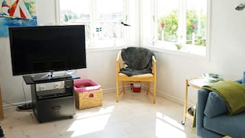 A living room with a flat-screen TV, a chair, a sofa, a small table, and a window with a view of buildings.